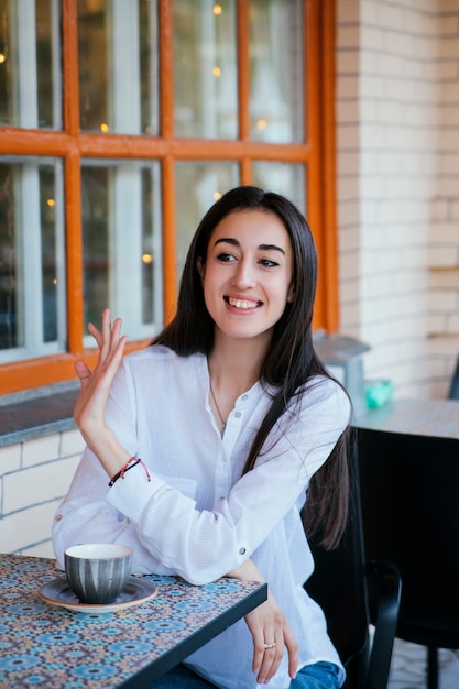 Una hermosa joven saluda a los transeúntes en una mesa de un café. Una niña bonita quiere hacer un pedido.