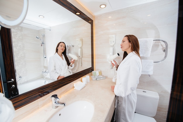 Una hermosa joven se seca el cabello con un secador de pelo en un hermoso baño blanco. Buenos días frescos en el hotel.