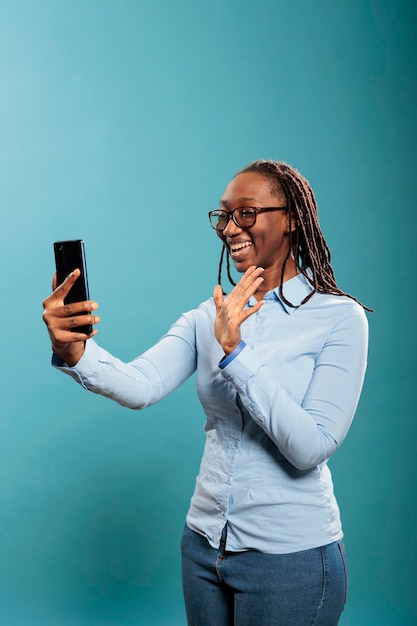 Hermosa joven segura de sí misma saludando a una amiga por videollamada en línea mientras está de pie en el fondo azul. Feliz sonriente mujer afroamericana con teléfono inteligente saludando en videoconferencia