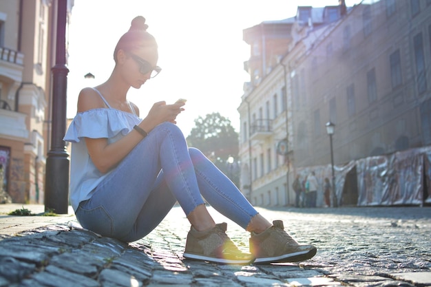 Hermosa joven sentada en un adoquín de una calle europea usando su smartphone