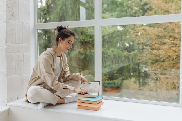 Hermosa joven sentada en el alféizar de la ventana sosteniendo y leyendo un libro