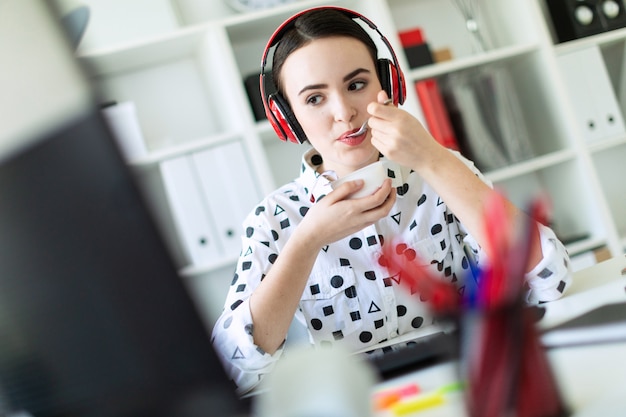 Hermosa joven sentada en auriculares en el escritorio en la oficina, comiendo yogurt y mirando el monitor.