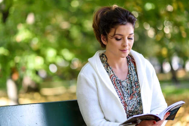 Hermosa joven sentada en un banco en el parque y leyendo un libro en un soleado día de otoño