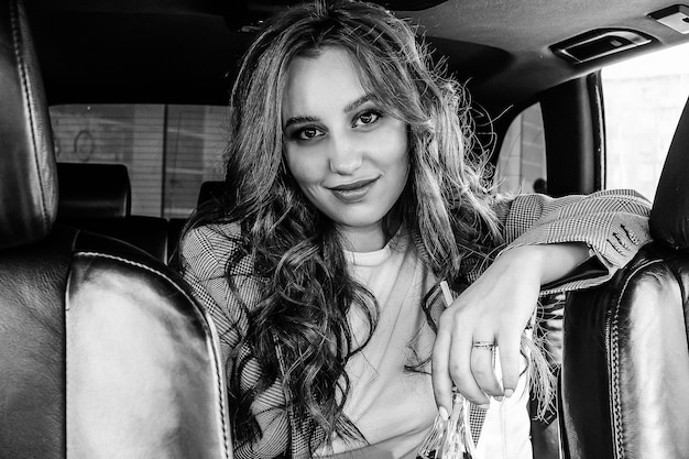 Hermosa joven sentada en un coche. Una chica elegante con un traje bebe limonada en el interior de un coche negro. Fotografía en blanco y negro.