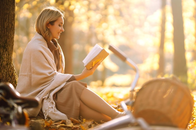 Hermosa joven sentada en hojas de otoño caídas en un parque leyendo un libro Relajación