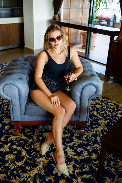 Hermosa joven sentada en un sillón suave en el vestíbulo del hotel con una copa de champán de vacaciones. Lleva gafas de sol y una sonrisa en su rostro. Concepto de vacaciones