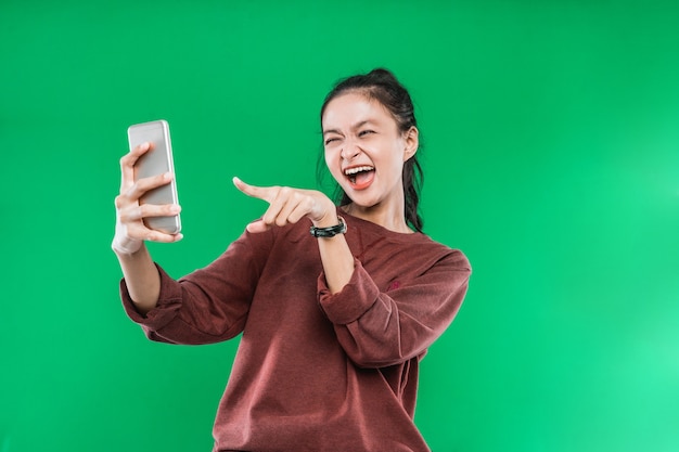 Hermosa joven siendo videollamada con alguien mientras apunta al teléfono con una expresión feliz sobre fondo verde