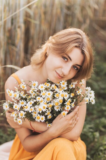 Una hermosa joven se sienta en un campo de trigo y tiene un ramo de margaritas en sus manos.