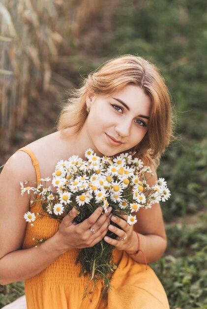 Una hermosa joven se sienta en un campo de trigo y tiene un ramo de margaritas en sus manos.