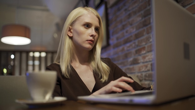 Una hermosa joven se sienta en Internet a través de una computadora portátil y hojea las noticias en un restaurante. Descanso en la gran ciudad, fin de semana. Primer plano, 4K UHD.