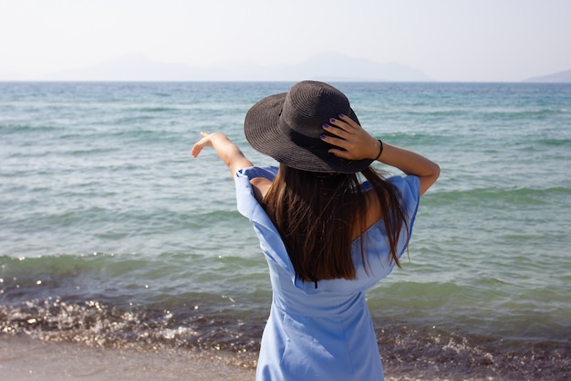 Hermosa joven con un sombrero mira el mar, muestra una mano