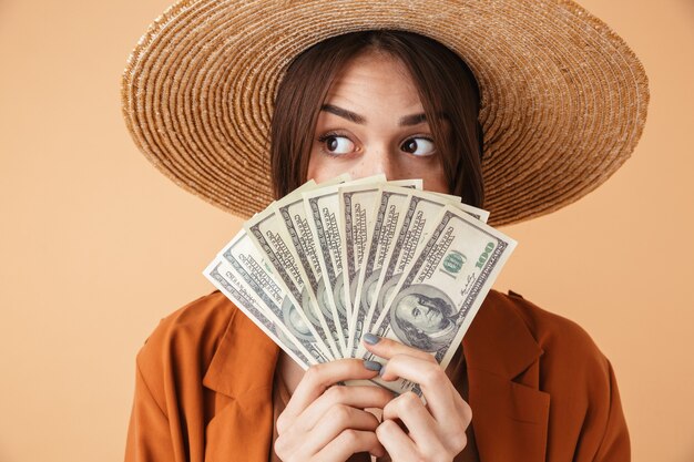 Hermosa joven con sombrero de paja y traje de verano que se encuentran aisladas sobre pared beige, mostrando billetes de dinero