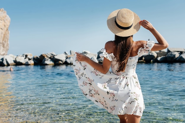 Una hermosa joven con un sombrero y un vestido ligero con la espalda camina a lo largo de la orilla del océano contra el fondo de enormes rocas en un día soleado Turismo y viajes de vacaciones