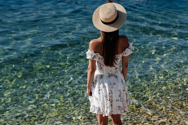 Una hermosa joven con un sombrero y un vestido ligero con la espalda camina a lo largo de la orilla del océano contra el fondo de enormes rocas en un día soleado Turismo y viajes de vacaciones