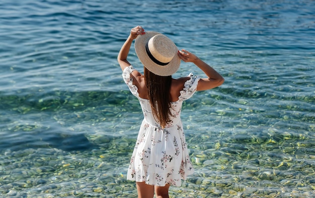 Una hermosa joven con un sombrero y un vestido ligero con la espalda camina a lo largo de la orilla del océano contra el fondo de enormes rocas en un día soleado Turismo y viajes de vacaciones