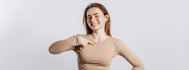 Hermosa joven sonriendo y apuntando con su dedo a sí misma sobre un fondo blanco aislado. Una mujer señala una idea, un lugar para publicidad. Morena positiva con un jersey beige.