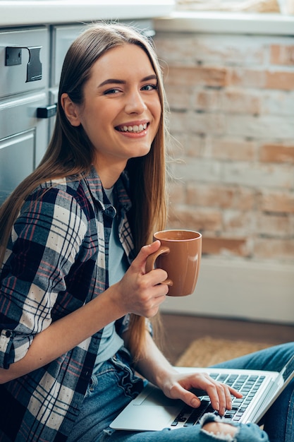 Hermosa joven sonriendo y bebiendo té mientras está sentado con un gadget en casa