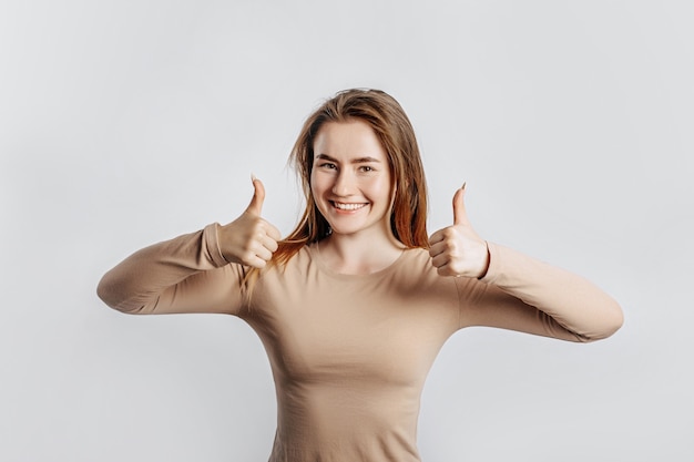 Foto hermosa joven sonriendo y muestra los pulgares para arriba gesto con las dos manos sobre un fondo blanco aislado. una mujer señala una idea, un lugar para publicidad. morena positiva con un jersey beige.