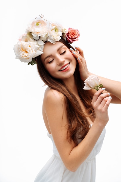 Hermosa joven sonriendo y posando con flores en la pared blanca en retrato de vestido blanco