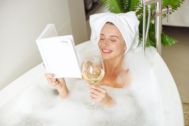Hermosa joven sonriente acostada en el baño con un libro de lectura de espuma y bebiendo vino