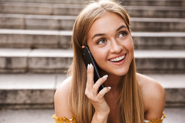 Hermosa joven sonriente hablando por teléfono móvil mientras está sentado en las escaleras al aire libre