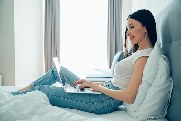 Hermosa joven sonriente en jeans y camiseta sentada en una cama en casa y sonriendo mientras usa una computadora portátil