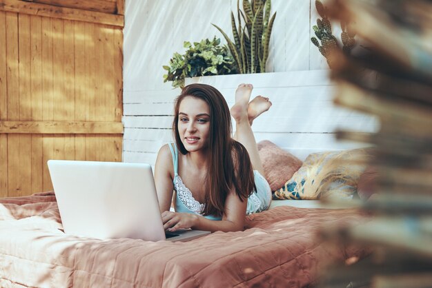 Hermosa joven sonriente con laptop mientras está acostado en la cama