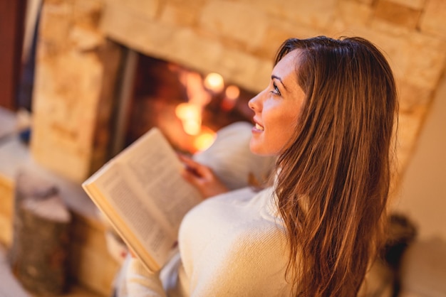 Hermosa joven sonriente leyendo un libro y disfrutando junto a la chimenea.