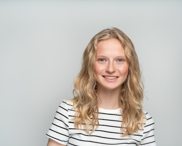 Hermosa joven sonriente a mujer rubia inteligente sin maquillaje en la pared blanca. Bonita mujer con pelo rizado en camiseta blanca