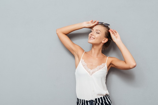 Hermosa joven sonriente con los ojos cerrados con gafas de sol y posando aislado en el fondo gris