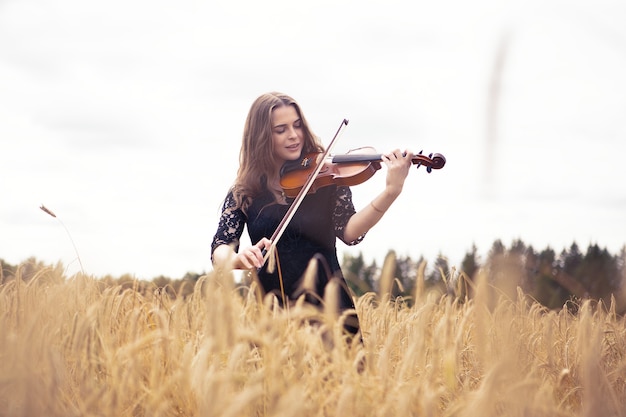 Hermosa joven sonriente de pie sobre un campo de trigo tocando el violín con entusiasmo