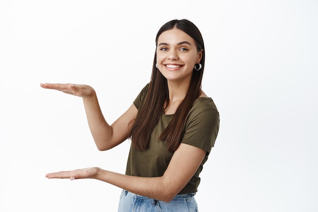 Foto hermosa joven sonriente presenta un producto que muestra un gesto de caja grande con un espacio de copia vacío para su logotipo o publicidad de fondo blanco