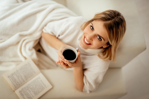 Hermosa joven sonriente relajándose en la cama con libro y café. Mirando a la cámara.