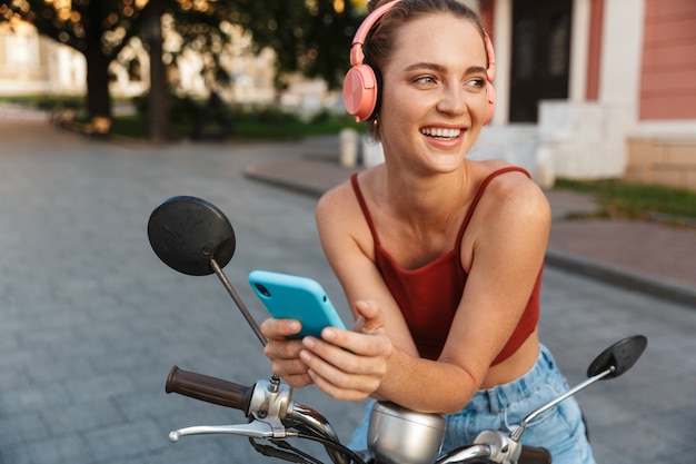 Hermosa joven sonriente con ropa casual de verano sentada en un scooter al aire libre en las calles de la ciudad, escuchando música con auriculares inalámbricos, sosteniendo el teléfono móvil