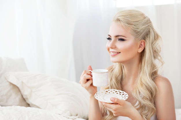 Hermosa joven sonriente sentada en la cama blanca, sosteniendo una taza de café