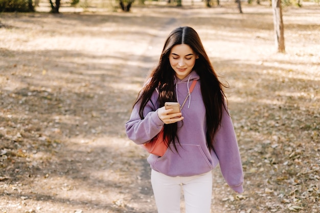 Hermosa joven sonriente con smartphone y caminar en el parque de otoño.