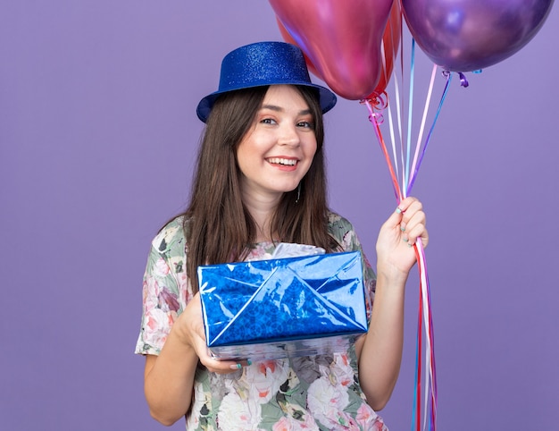 Hermosa joven sonriente con sombrero de fiesta sosteniendo globos con caja de regalo aislada en la pared azul