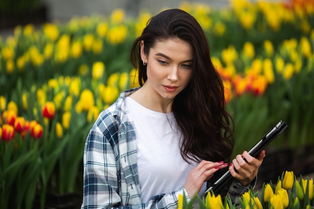 Hermosa joven sonriente con tableta, trabajador con flores en invernadero.