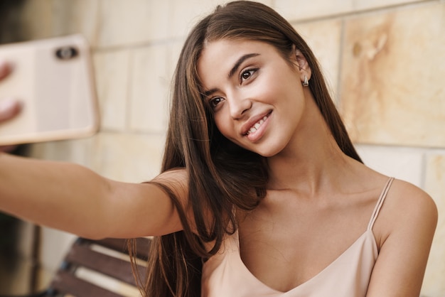 Hermosa joven sonriente con vestido de verano sentado en un banco al aire libre, tomando un selfie