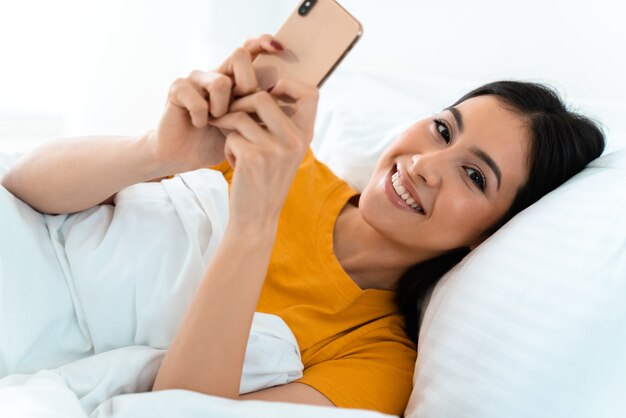 una hermosa joven sonriente yace en la cama en el interior de su casa mediante teléfono móvil.
