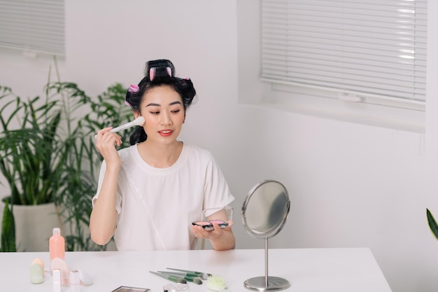 Hermosa joven se sonroja en la cara usando un cepillo de maquillaje mientras se sienta en casa en una habitación en una mesa