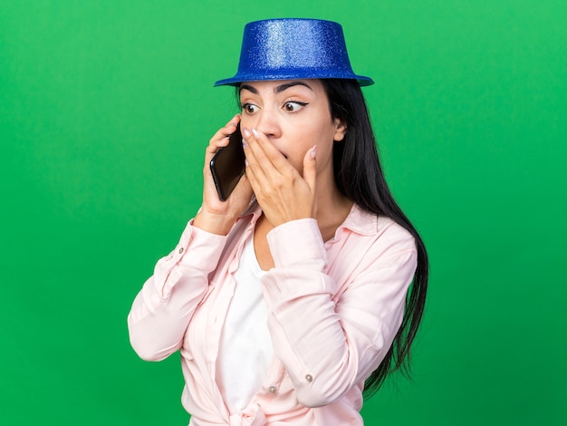 Hermosa joven sorprendida con gorro de fiesta habla por teléfono con la boca cubierta con la mano