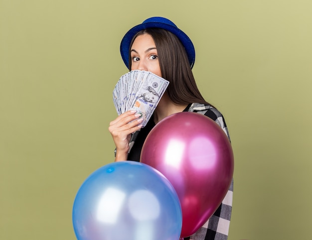 Hermosa joven sorprendida con sombrero azul de pie detrás de globos con la cara cubierta con dinero en efectivo aislado en la pared verde oliva