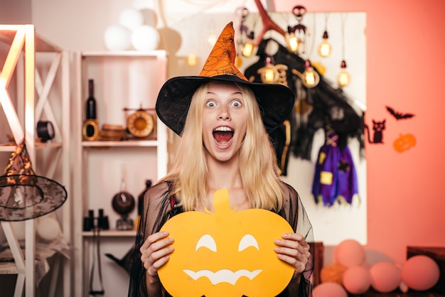 Hermosa joven sorprendida con sombrero de brujas y traje con calabaza. Linterna de cabeza de calabaza. Truco o trato. Bastante joven bruja rubia volando en la escoba, aislada sobre fondo naranja.