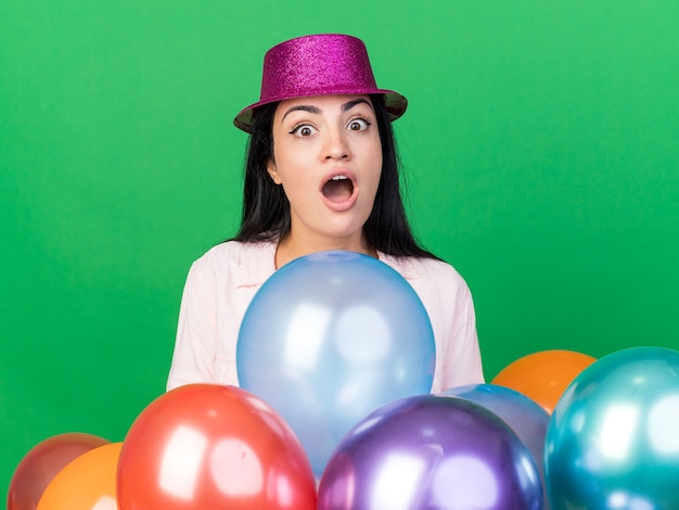 Hermosa joven sorprendida con sombrero de fiesta de pie detrás de globos