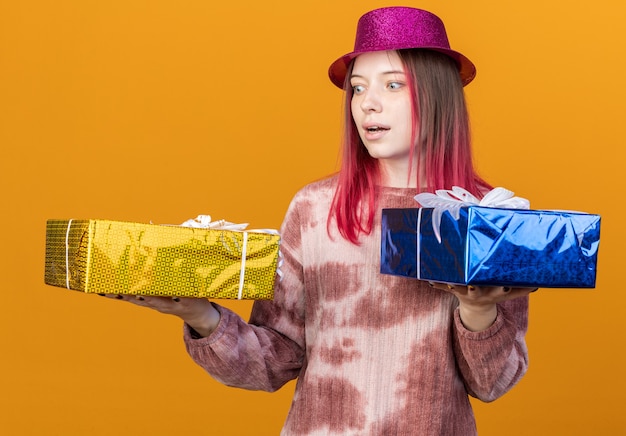 Hermosa joven sorprendida con sombrero de fiesta sosteniendo y mirando cajas de regalo