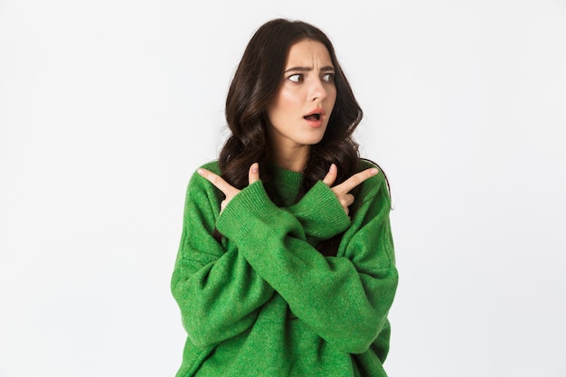 Hermosa joven sorprendida vestida con suéter verde posando aislado en la pared blanca apuntando.