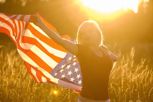 Hermosa joven sosteniendo una bandera americana en el viento en un campo de centeno. Paisaje de verano contra el cielo azul. Orientación horizontal.