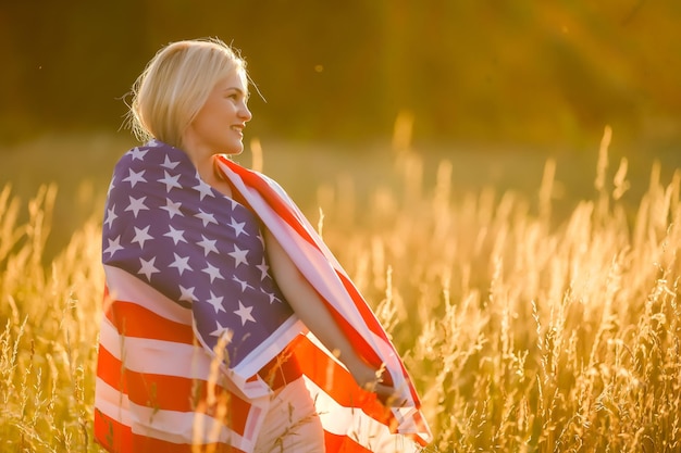 Hermosa joven sosteniendo una bandera americana en el viento en un campo de centeno. Paisaje de verano contra el cielo azul. Orientación horizontal.