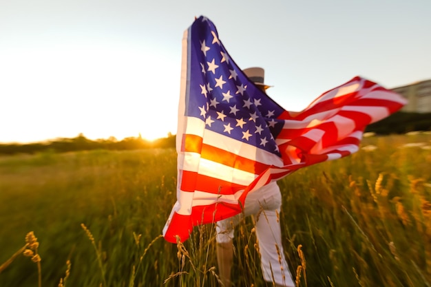 Hermosa joven sosteniendo una bandera americana en el viento en un campo de centeno. Paisaje de verano contra el cielo azul. Orientación horizontal.